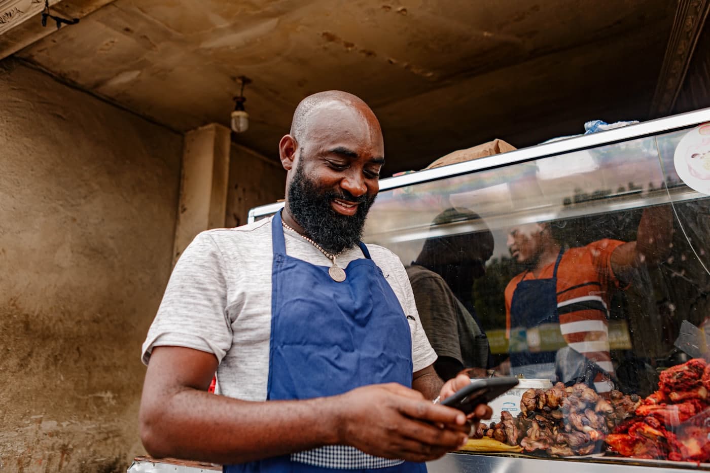 El dueño de un negocio con delantal sonriendo frente a su teléfono, detrás del mostrador de su local de comida.